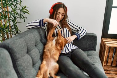 Young hispanic woman listening to music sitting on sofa with dog at home