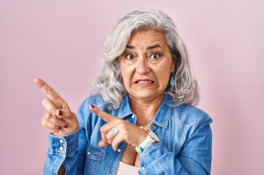 Middle age woman with grey hair standing over pink background pointing aside worried and nervous with both hands, concerned and surprised expression 