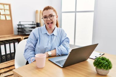 Ofiste çalışan genç kızıl saçlı kadın bilgisayarını kullanıyor. Dilini komik bir ifadeyle dışarı çıkarıyor. duygu kavramı. 