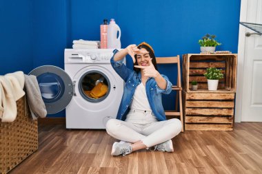 Hispanic woman doing laundry sitting on the floor smiling making frame with hands and fingers with happy face. creativity and photography concept. 
