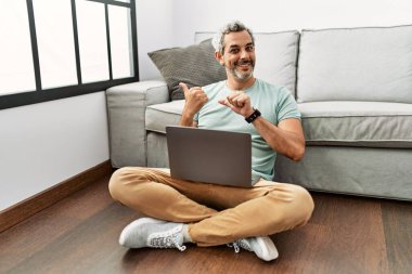 Orta yaşlı İspanyol bir adam oturma odasında dizüstü bilgisayar kullanıyor. Arka tarafı işaret ediyor. El ve baş parmakları havada, kendinden emin bir şekilde. 