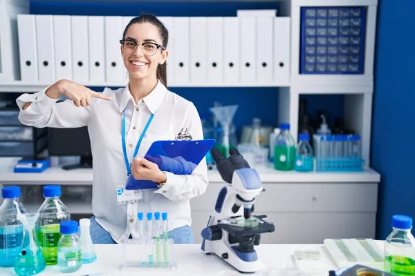 Young brunette woman working at scientist laboratory looking confident with smile on face, pointing oneself with fingers proud and happy.
