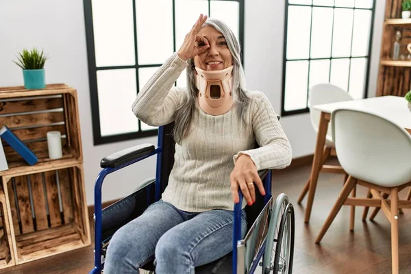 Middle age grey-haired woman sitting on wheelchair with collar at home smiling happy doing ok sign with hand on eye looking through fingers 