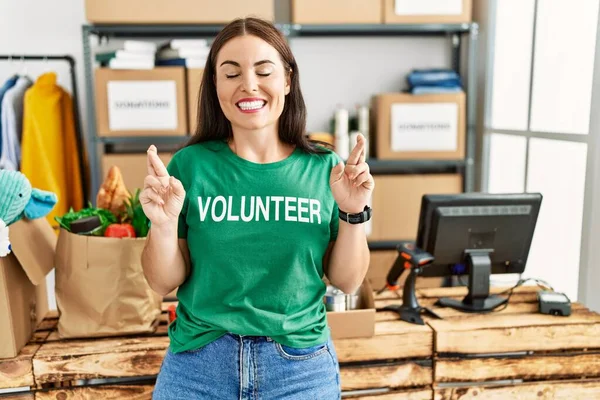 Jovem Morena Vestindo Shirt Voluntária Doações Gesto Dedo Cruzado Sorrindo — Fotografia de Stock