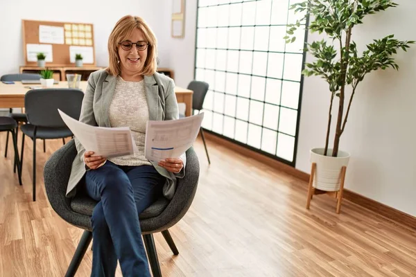 Middle age businesswoman smiling happy reading paperwork sitting at the office.
