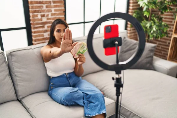 Young hispanic woman recording tutorial with smartphone at home with open hand doing stop sign with serious and confident expression, defense gesture