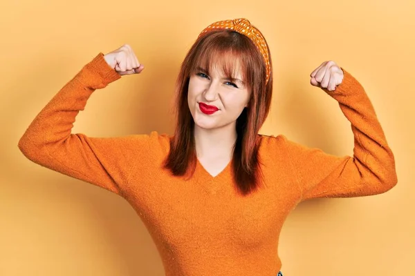 红头发的年轻女人穿着休闲的橙色毛衣 露出胳膊肌肉 露出得意的笑容 健身概念 — 图库照片