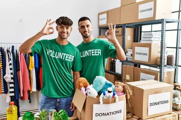 Young gay couple wearing volunteer t shirt at donations stand smiling positive doing ok sign with hand and fingers. successful expression.