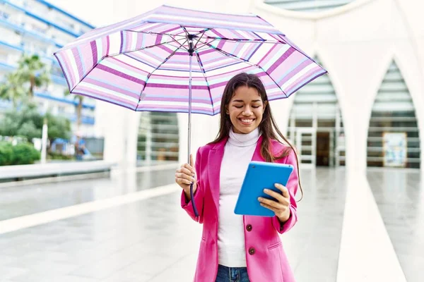 年轻的拉丁女人在街上拿着雨伞用触摸板 — 图库照片