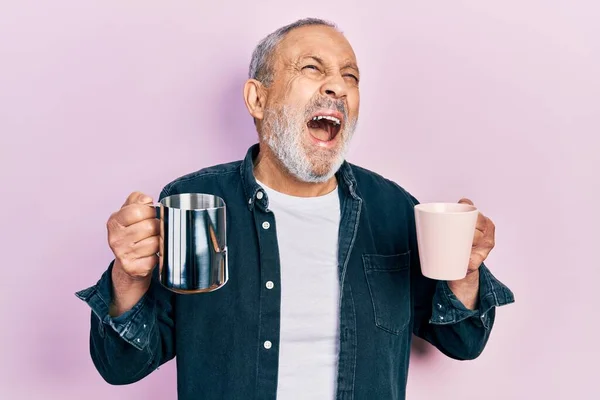 Handsome senior man with beard holding coffee and milk angry and mad screaming frustrated and furious, shouting with anger looking up.