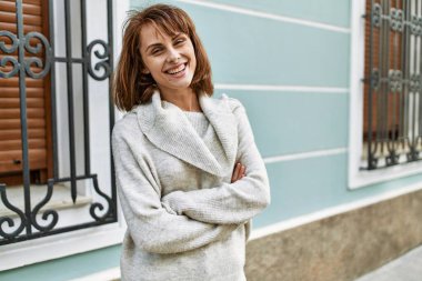 Genç beyaz kız gülümsüyor. Kollarını kavuşturup şehirde mutlu bir şekilde duruyor..