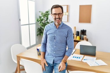 Middle age hispanic man with beard wearing business clothes at the office winking looking at the camera with sexy expression, cheerful and happy face. 