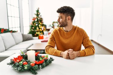 Arap genç adam Noel ağacının yanında oturmuş yan yan bakıyor, rahat bir görünüm takınıyor kendine güvenen bir gülümsemeyle doğal bir yüzle.. 