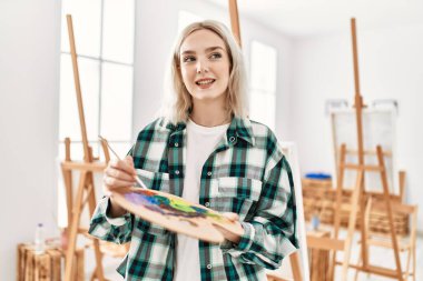 Resim stüdyosunda fırça ve palet tutarak mutlu gülümseyen genç sanatçı kız..