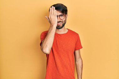 Gündelik kıyafetler ve gözlük takan genç İspanyol bir adam bir gözünü eliyle kapatıyor, yüzünde kendine güvenen bir gülümseme ve sürpriz bir duygu var.. 