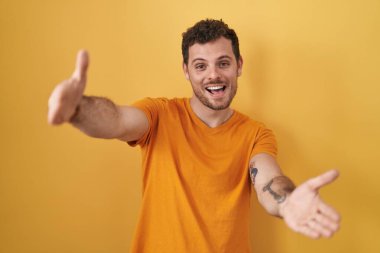 Sarı arka planda dikilen İspanyol genç adam kameraya bakıp sarılmak için kollarını açmış gülümsüyor. Mutluluğu kucaklayan neşeli bir ifade.. 