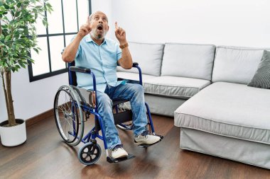 Handsome senior man sitting on wheelchair at the living room amazed and surprised looking up and pointing with fingers and raised arms. 
