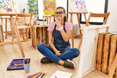 Sanat stüdyosundaki genç esmer kadın yerde oturmuş kameraya kendinden emin bir şekilde bakıyor kollarını kavuşturup ve çenesini kaldırarak gülümsüyor. Olumlu düşünmek. 