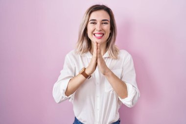 Young beautiful woman standing over pink background praying with hands together asking for forgiveness smiling confident. 