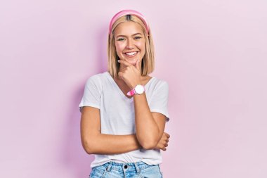 Beautiful blonde woman wearing casual white t shirt looking confident at the camera smiling with crossed arms and hand raised on chin. thinking positive. 
