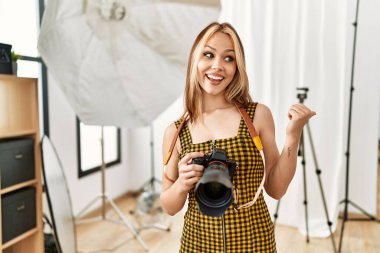 Young caucasian photographer girl holding professional camera at photography studio pointing thumb up to the side smiling happy with open mouth 