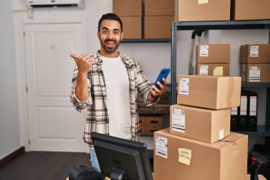 Sakallı, küçük işletme ekommerinde çalışan akıllı telefonlu, ağzı açık gülümseyen genç İspanyol bir adam. 