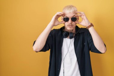 Young caucasian man wearing sunglasses standing over yellow background trying to open eyes with fingers, sleepy and tired for morning fatigue 