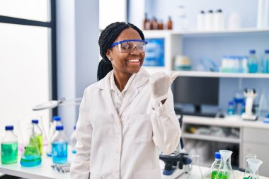 African american woman working at scientist laboratory pointing thumb up to the side smiling happy with open mouth 