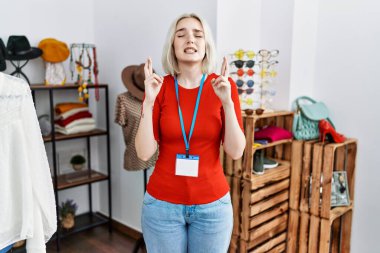 Young caucasian woman working as manager at retail boutique gesturing finger crossed smiling with hope and eyes closed. luck and superstitious concept. 