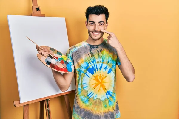画家のイーゼルスタンドでパレットで絵を描く若いヒスパニック系の男が顔や鼻に手指で指して立って 陽気に笑顔 美の概念 — ストック写真