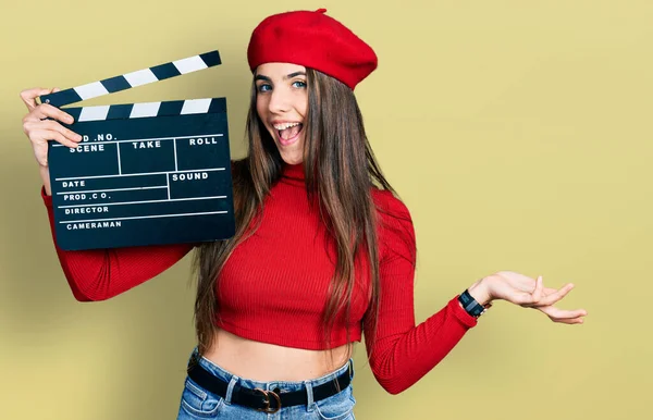 Young brunette teenager holding video film clapboard celebrating achievement with happy smile and winner expression with raised hand