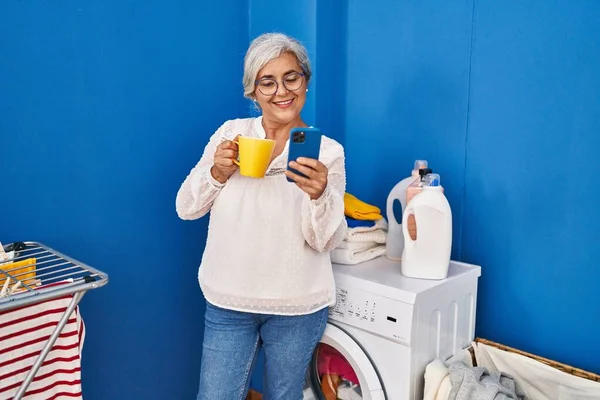 Middle Age Woman Using Smartphone Drinking Coffee Waiting Washing Machine — Stock Photo, Image