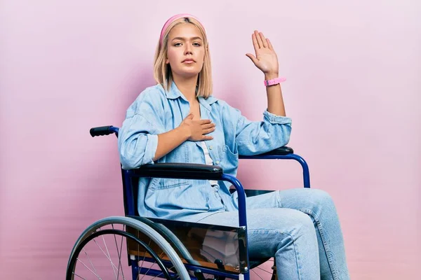 Beautiful blonde woman sitting on wheelchair swearing with hand on chest and open palm, making a loyalty promise oath