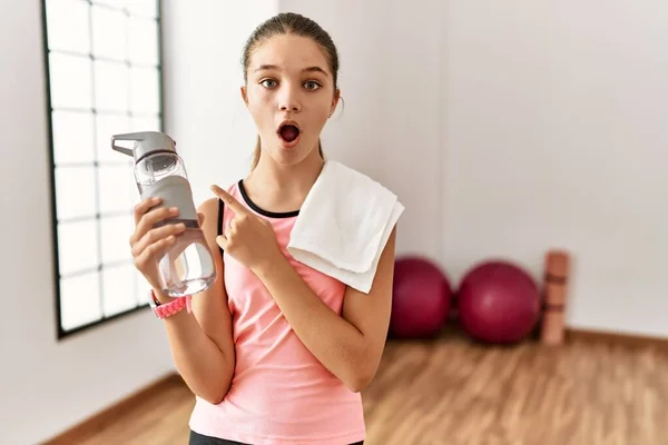 Young Brunette Teenager Wearing Sportswear Holding Water Bottle Surprised Pointing — Zdjęcie stockowe