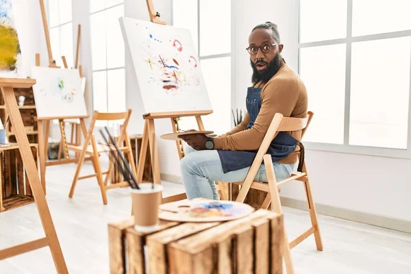 African american artist man painting on canvas at art studio scared and amazed with open mouth for surprise, disbelief face