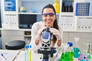 Young beautiful hispanic woman scientist smiling confident using microscope at laboratory