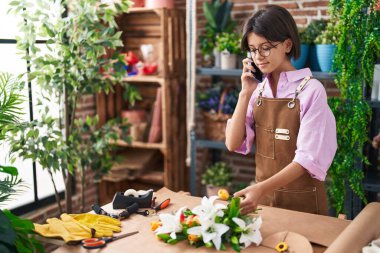Adorable hispanic girl florist talking on smartphone make bouquet of flowers at flower shop