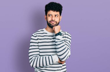 Sakallı, sıradan çizgili süveter giyen, eli çenesinde soru soran genç bir Arap. Düşünceli bir ifade. Gülümseyen ve düşünceli bir yüz. şüphe kavramı. 