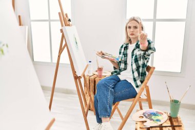 Young artist woman painting on canvas at art studio pointing with finger up and angry expression, showing no gesture 