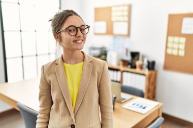 Young brunette teenager wearing business style at office looking away to side with smile on face, natural expression. laughing confident. 