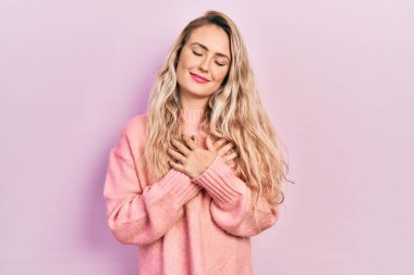 Beautiful young blonde woman wearing pink sweater smiling with hands on chest with closed eyes and grateful gesture on face. health concept. 