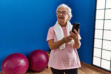 Spor kıyafetleri giyen yaşlı gri saçlı kadın spor merkezinde akıllı telefon kullanıyor.