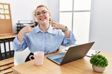 Ofiste çalışan genç kızıl saçlı kadın bilgisayarlı dizüstü bilgisayar kullanıyor gülümsüyor ve parmakları ve ağzı ile işaret ediyor. diş sağlığı kavramı. 