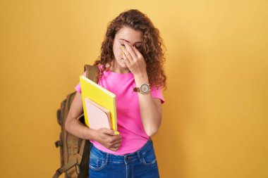 Öğrenci çantası takan, kitap tutan, burnu ve gözleri ovuşturmaktan yorgun düşmüş, yorgun ve başı ağrıyan beyaz bir kadın. Stres ve hüsran kavramı. 
