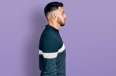 Sakallı, kışlık süveter giyen genç İspanyol bir adam. Yan tarafa bakıyor, rahat poz veriyor, kendine güvenen bir gülümsemesi var.. 