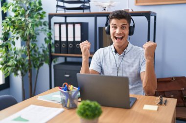 Young hispanic man working at the office wearing headphones celebrating surprised and amazed for success with arms raised and open eyes. winner concept. 