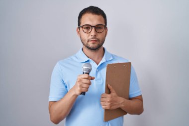 Hispanic man holding reporter microphone and clipboard relaxed with serious expression on face. simple and natural looking at the camera. 