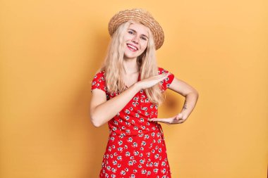 Beautiful caucasian woman with blond hair wearing summer hat gesturing with hands showing big and large size sign, measure symbol. smiling looking at the camera. measuring concept. 