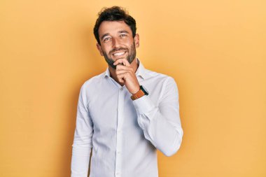 Sakallı, sıradan beyaz tişörtlü yakışıklı, kameraya kollarını kavuşturup çenesine dokunarak gülümseyen. Olumlu düşünmek. 