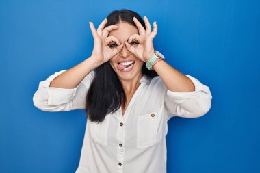 Mavi arka planda duran genç İspanyol kadın dürbün gibi jest yapıyor, gözleri parmaklarından bakıyor. çılgın ifade. 